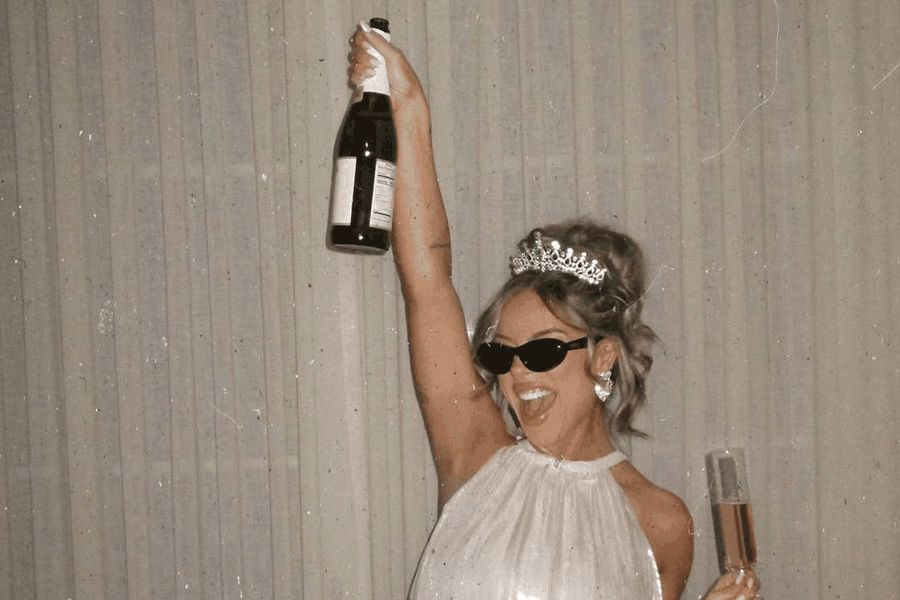 A bride to be in a feather-trimmed dress holding a champagne bottle above her head
