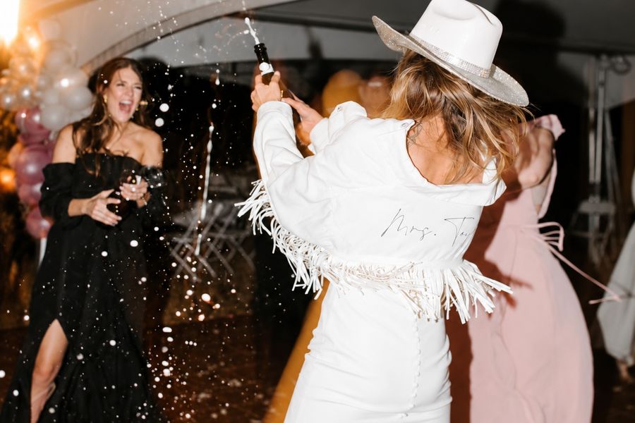Friends spraying champagne, the bride to be in white with a wide-brimmed hat
