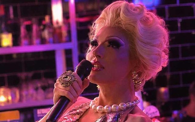 A drag performer in sequins and pearls singing into a microphone under pink bar lights
