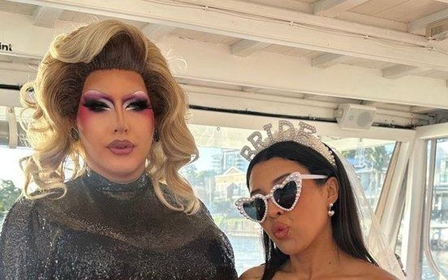 A drag queen in glitter with the bride to be in white and heart sunglasses