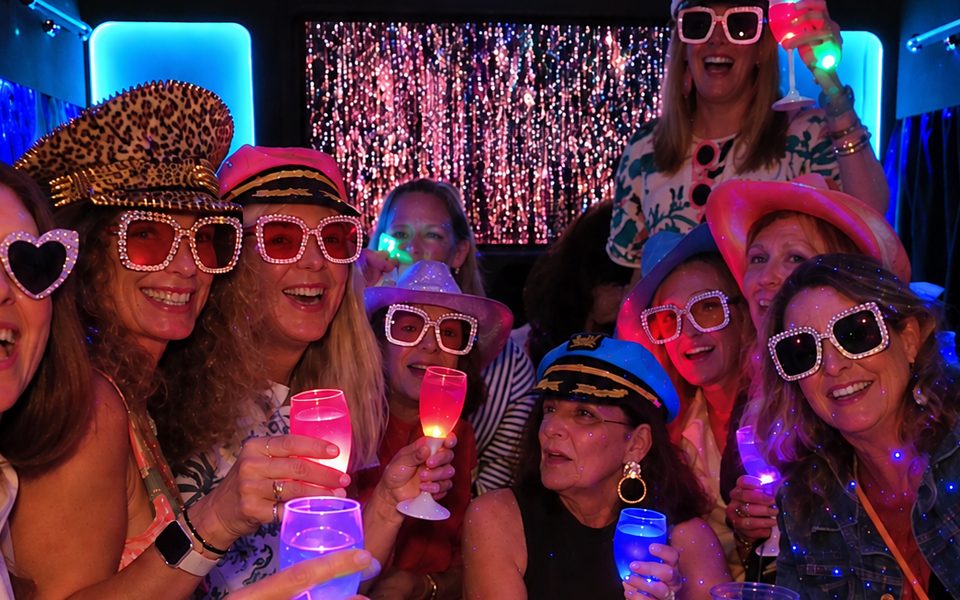 A hens group in party hats and sparkly sunglasses raising glowing glasses on a party bus