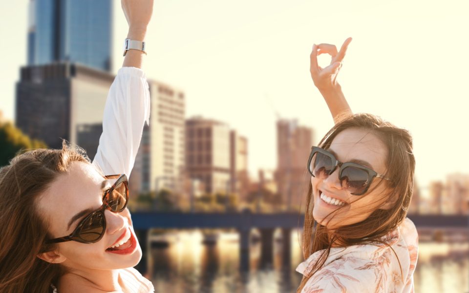 Two friends in sunglasses laughing in golden light with a city skyline and river behind them