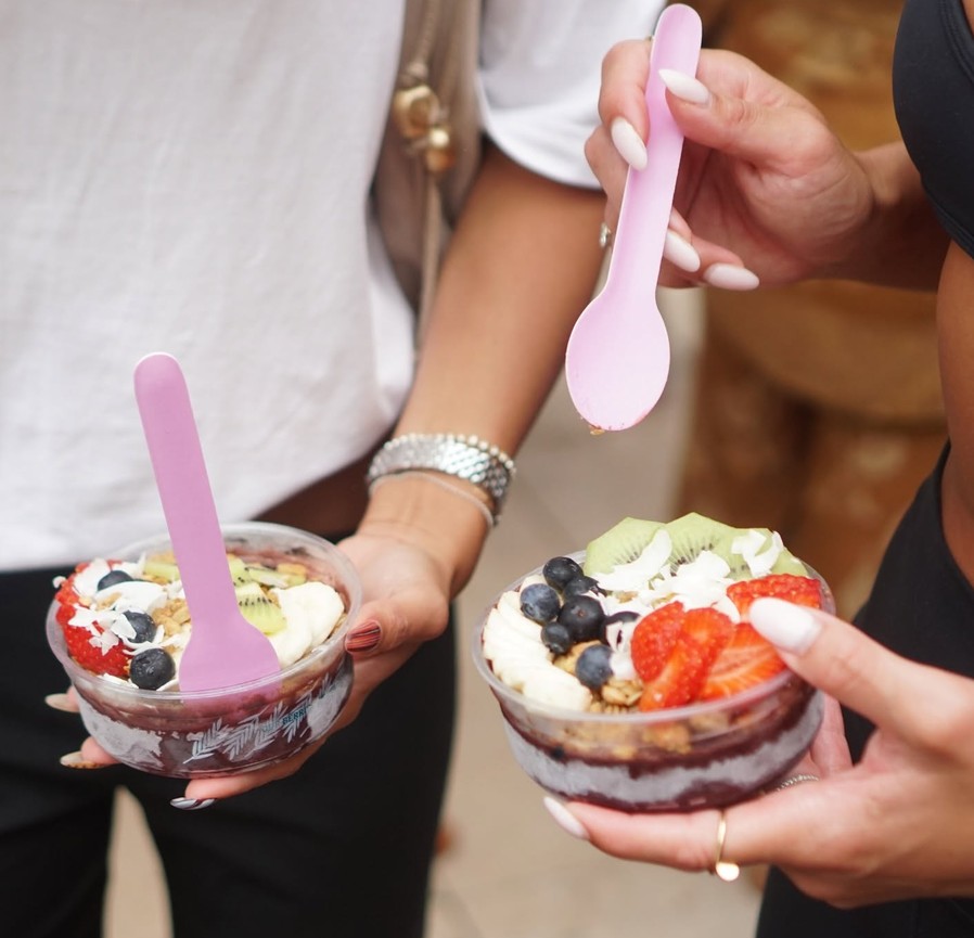 Two friends holding acai bowls topped with strawberries, kiwi fruit and blueberries, with pink spoons