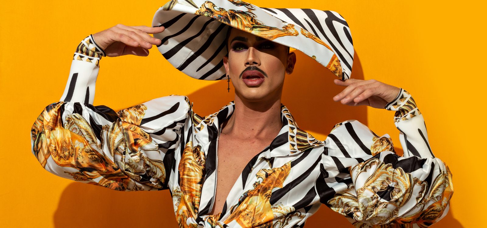 Drag performer in a zebra and gold print outfit and wide brimmed hat against an orange background