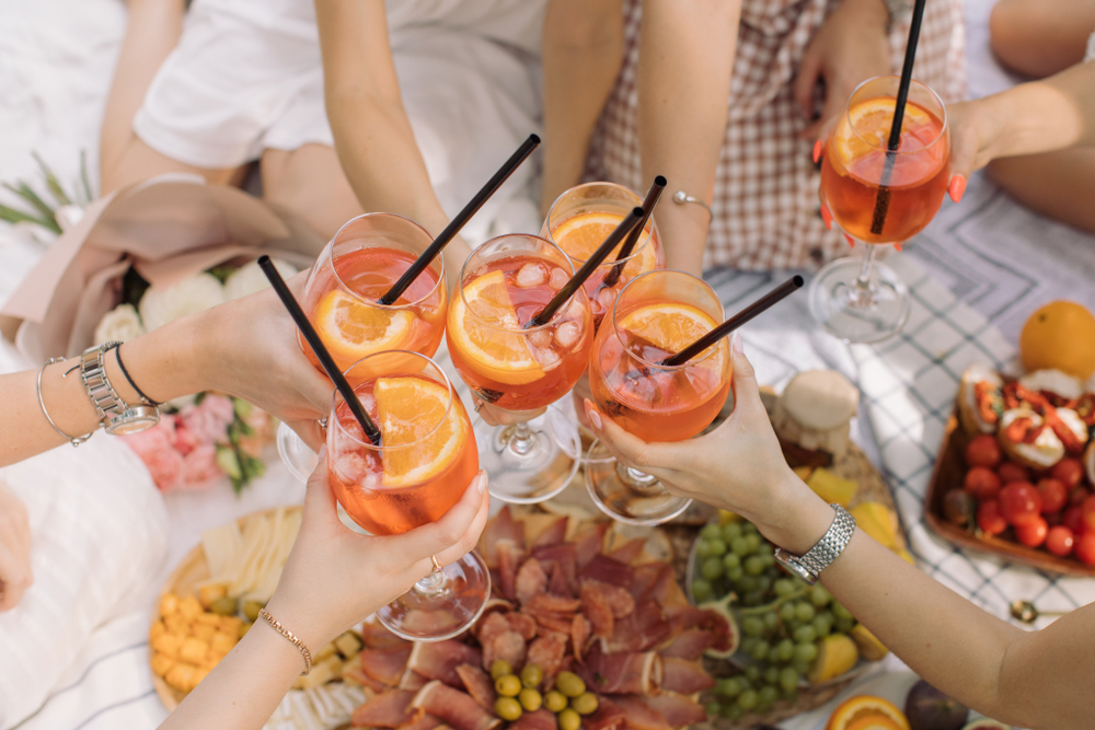 Friends raising glasses of orange spritz over a picnic spread