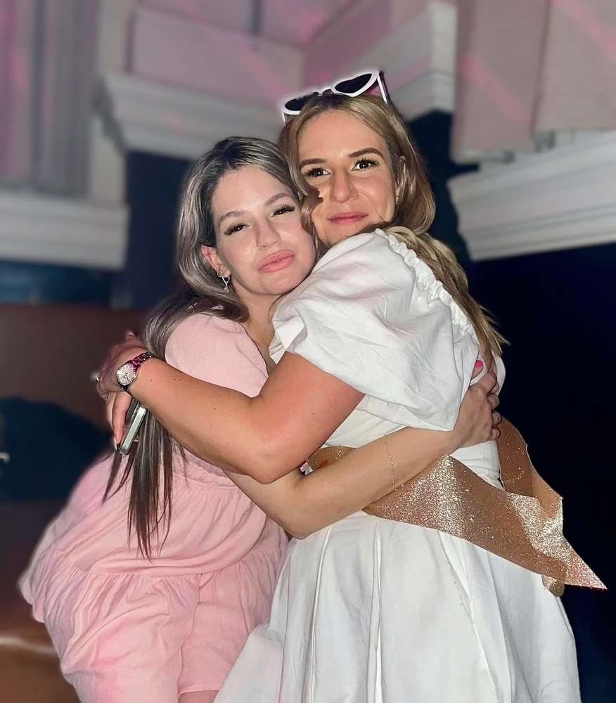 Two friends hugging tightly, one in a white dress with a sparkly sash