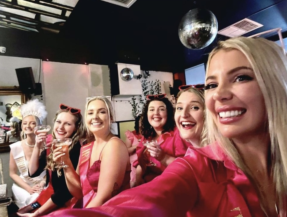 A hens group in pink raising cocktails for a selfie under a mirror ball, the bride to be in white with a feather headpiece