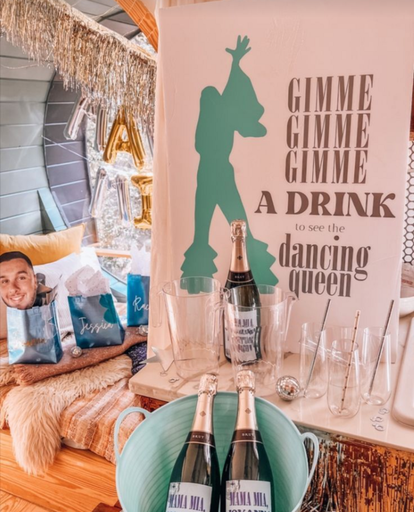 A sign reading gimme gimme gimme a drink, dancing queen, above bottles of bubbly