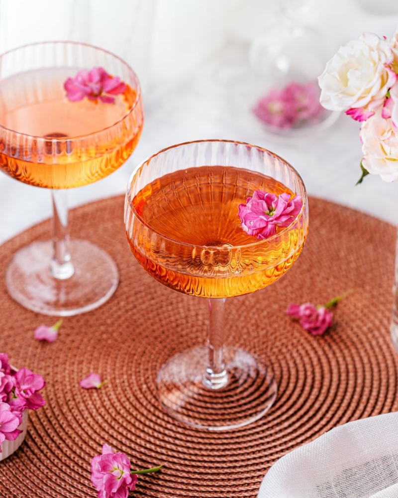 An orange cocktail in a ribbed coupe on a woven placemat scattered with pink petals
