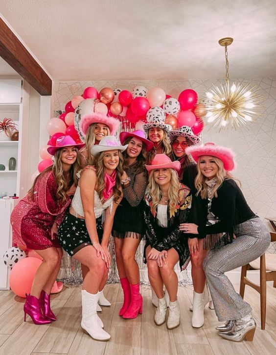 A group of friends in pink cowboy hats and boots posing in front of pink and red balloons