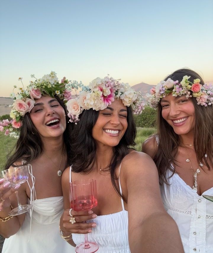 Three friends in flower crowns and white dresses taking a selfie at sunset