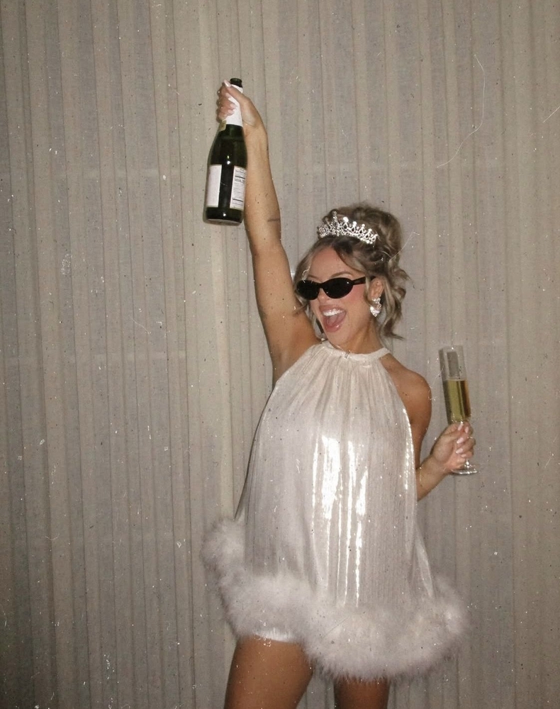 A bride in a white mini with feather trim, a tiara and heart sunglasses, holding a bottle of champagne up high