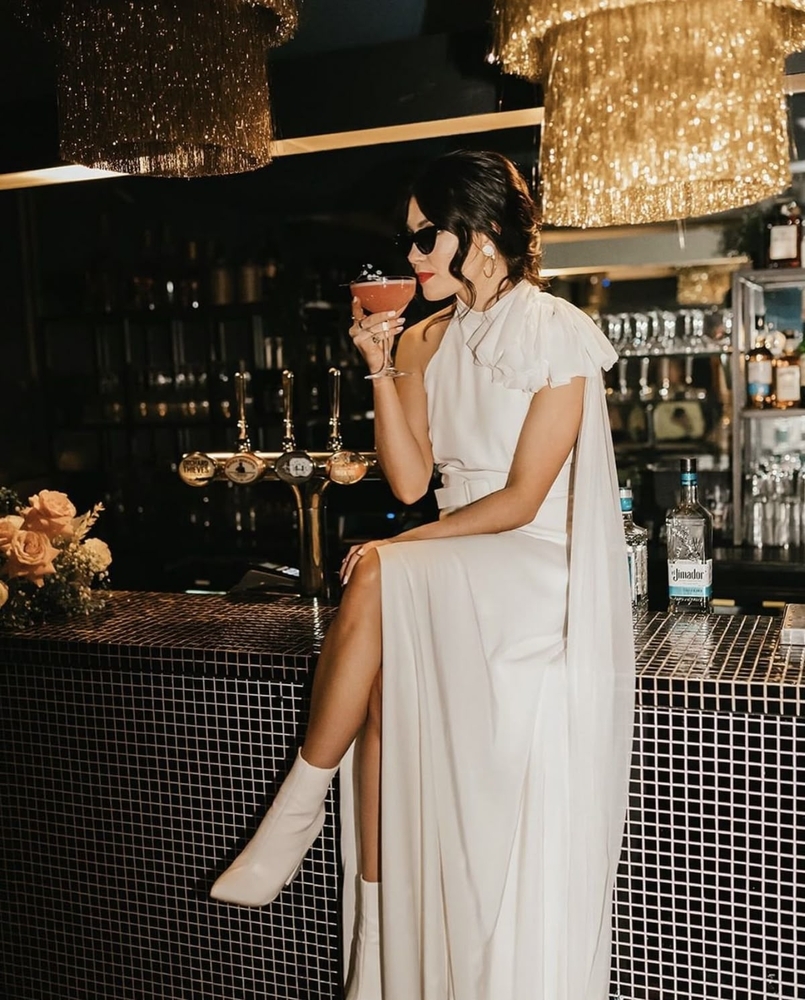 A bride in a long white dress and white boots sitting on a bar with a cocktail and sunglasses