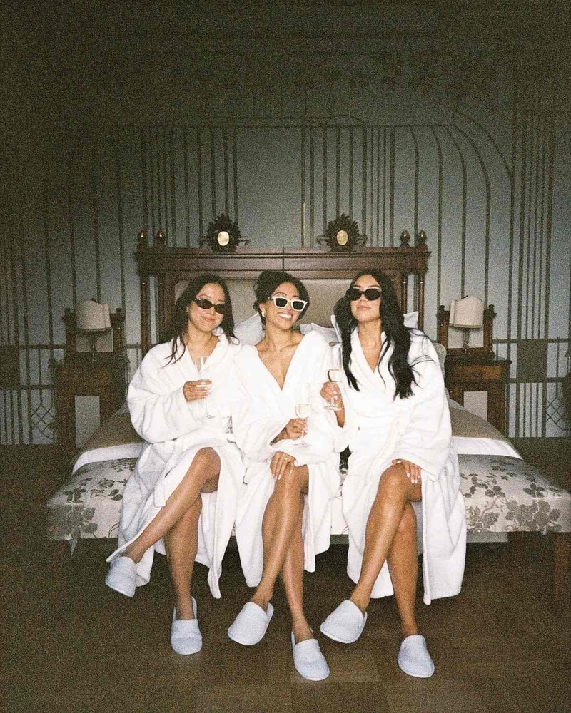 Three friends in fluffy white robes, slippers and sunglasses sitting on the end of a bed