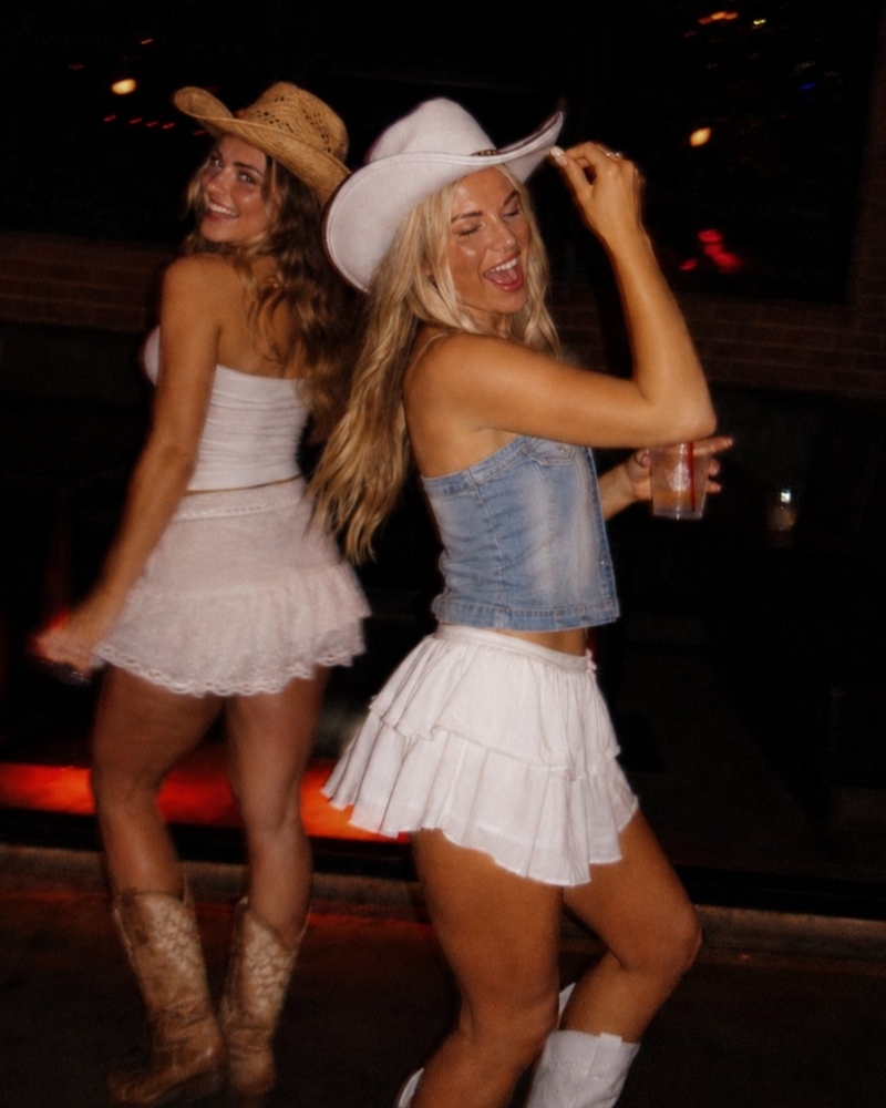 Two friends in cowboy hats and white skirts dancing together at night