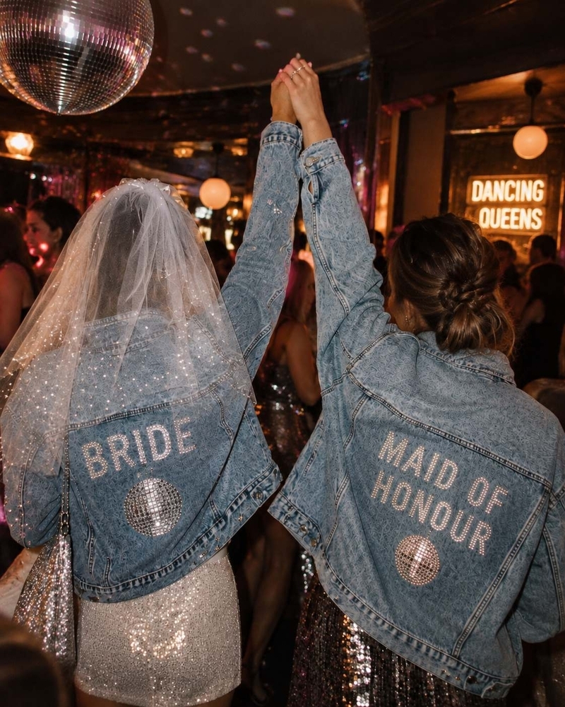 The backs of two denim jackets reading BRIDE and MAID OF HONOUR in rhinestones, hands joined in the air under a disco ball