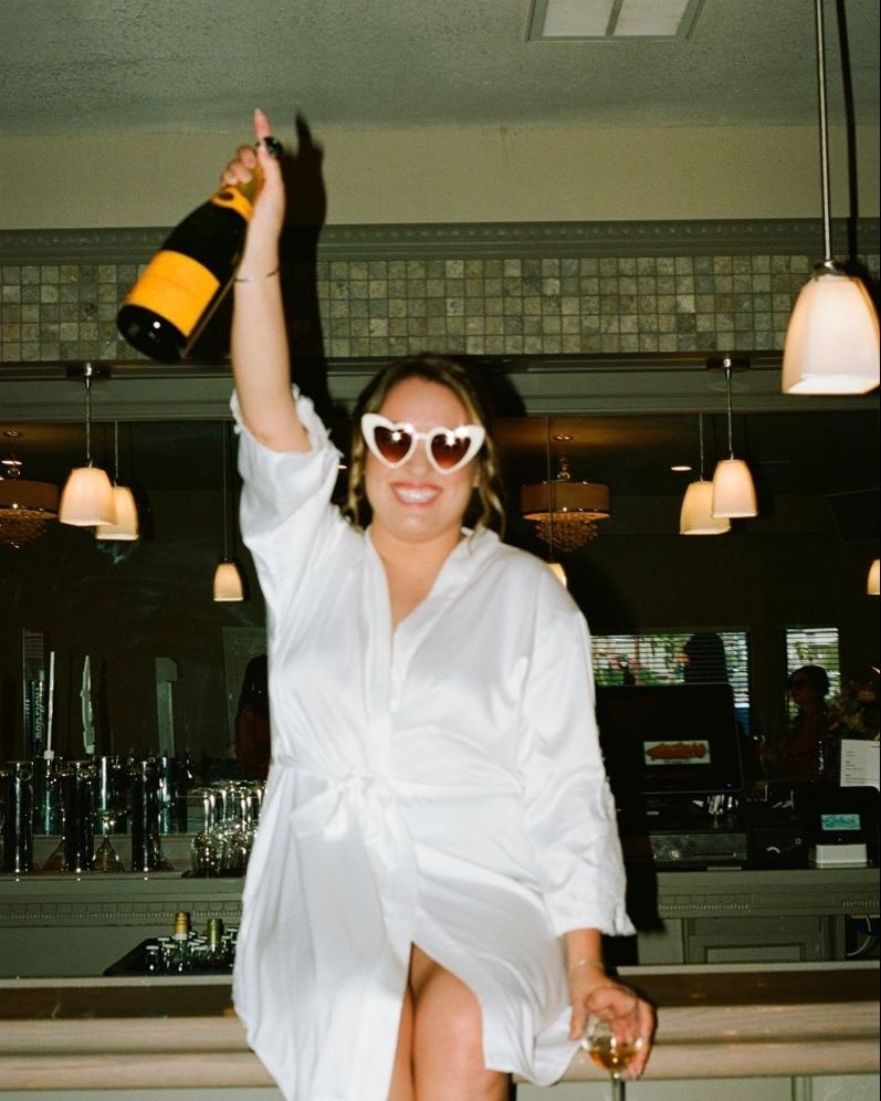 A hen in a white robe and heart sunglasses raising a bottle of champagne
