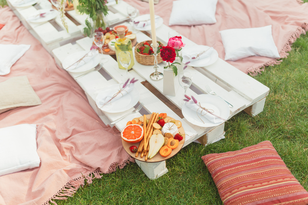 A low picnic table set on the grass with pink rugs, cushions, flowers and a grazing board