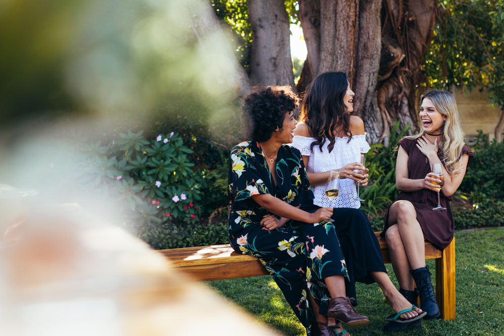Three friends laughing on a garden bench with glasses of white wine under the trees