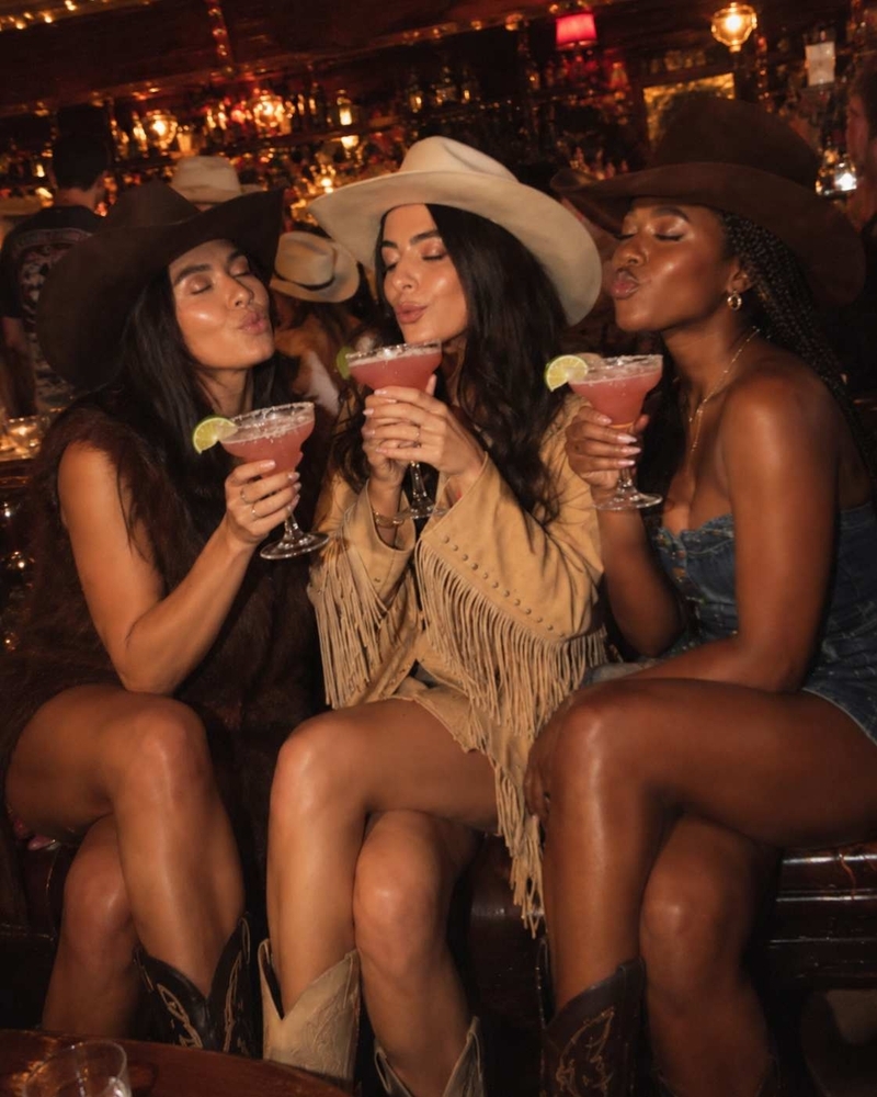 Three friends in cowboy hats, fringe and denim clinking pink margaritas with salted rims in a dim bar