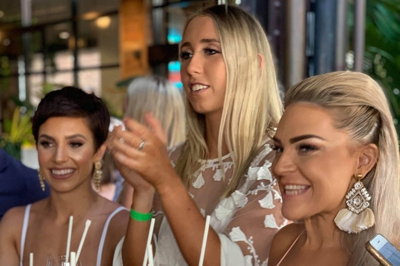 Friends laughing and clapping at a waterfront table lined with orange spritz cocktails