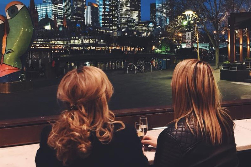 Two friends with glasses of bubbly looking across the river at a lit city skyline