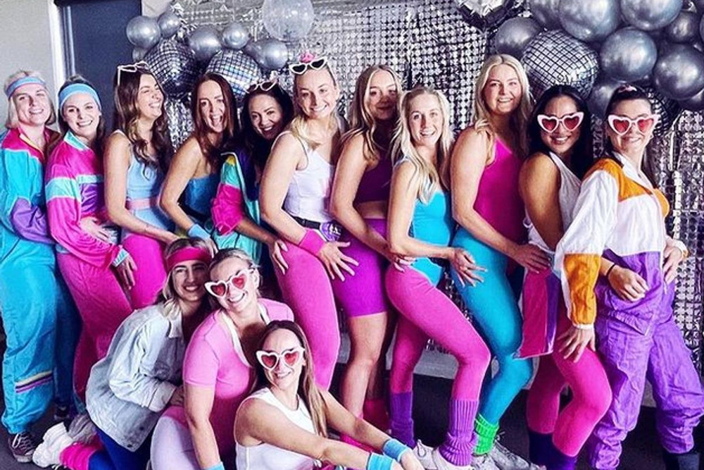 A hens group in bright retro tracksuits and heart sunglasses in front of silver balloons