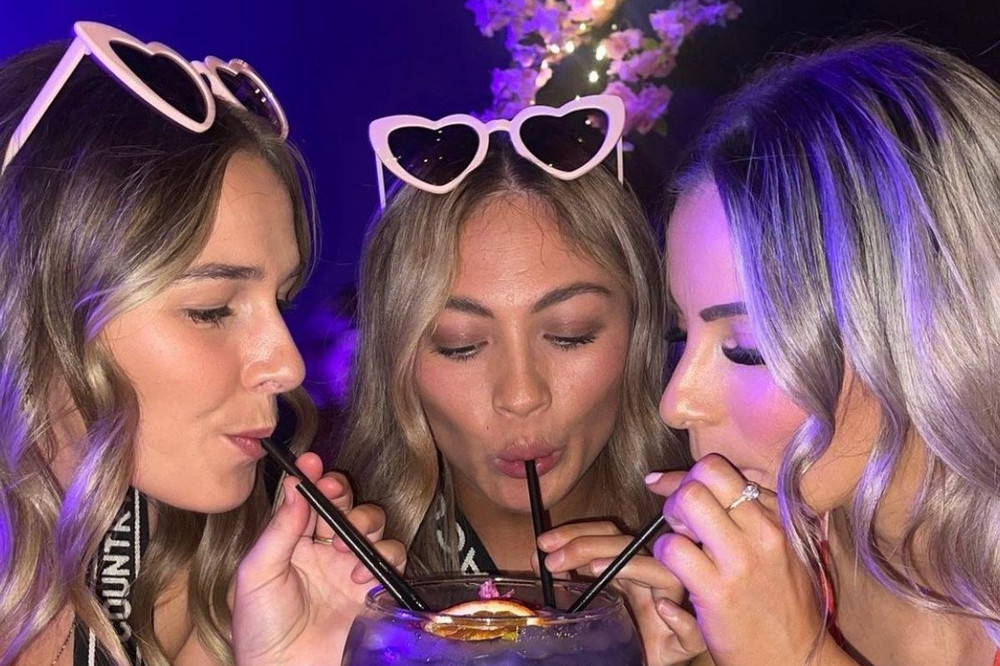 Three friends in heart sunglasses sharing one cocktail through straws under purple light