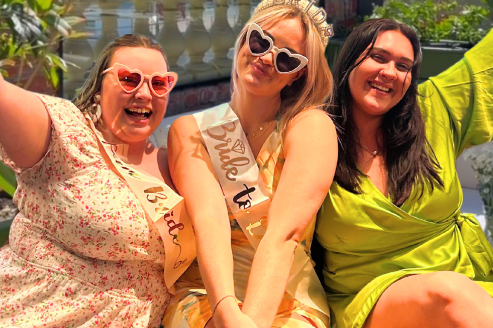 The bride in a sash and heart sunglasses laughing with two friends on a sunny city rooftop
