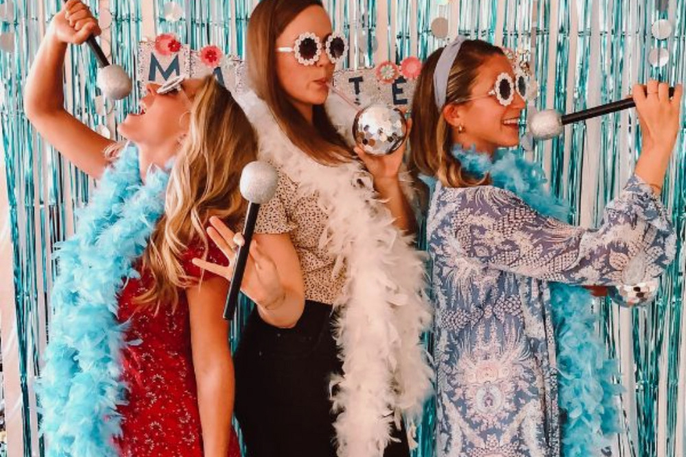 Three friends in heart sunglasses and feather boas singing into disco ball microphones against blue tinsel