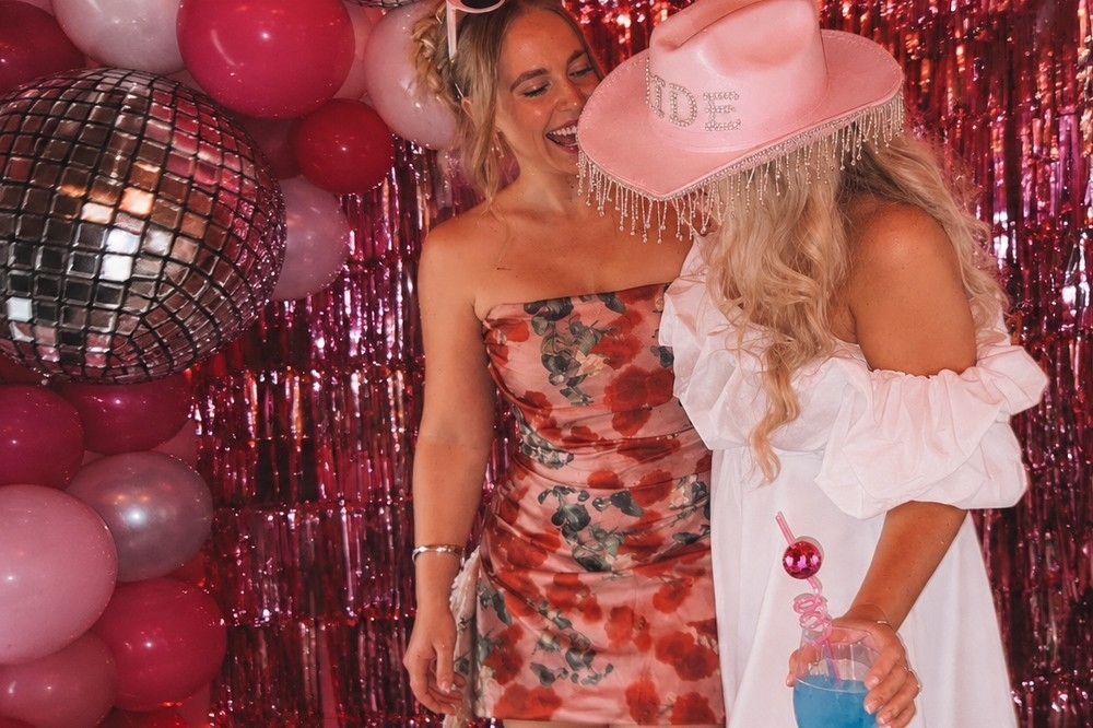A bride in a pink cowgirl hat with rhinestone fringe laughing with a friend in front of a pink tinsel curtain, a disco ball and pink balloons