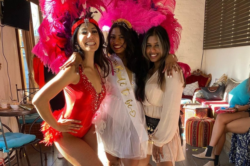 The bride in her sash beside our burlesque host in a red costume and pink feather headdress