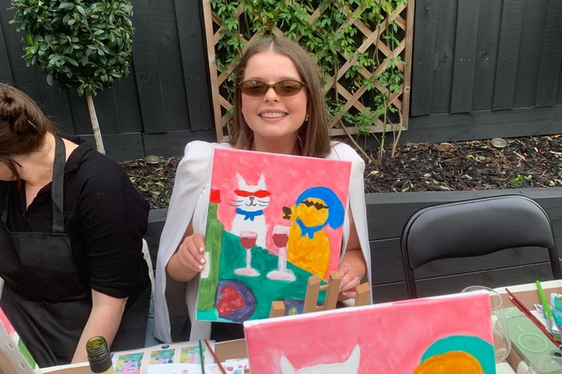 A guest in sunglasses holding up her painting of a cat and a dog in sunglasses with two glasses of red