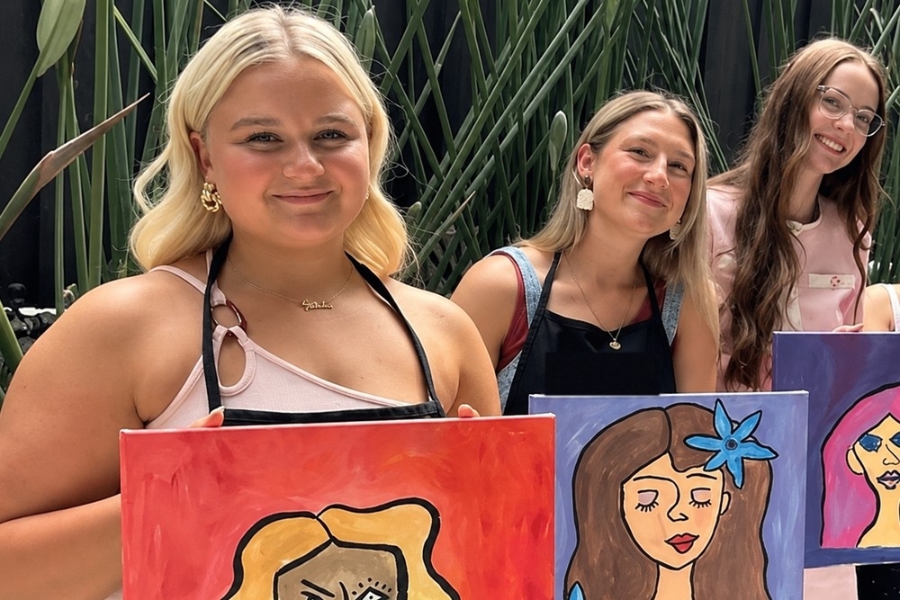Friends at a painting class holding up their finished portraits, one wearing an apron