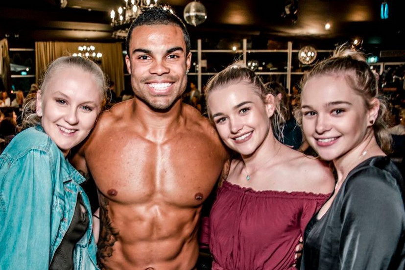 Three smiling friends posing with our shirtless waiter at a busy bar