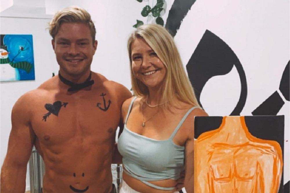 A shirtless waiter in a bow tie beside a guest holding up her painting of him