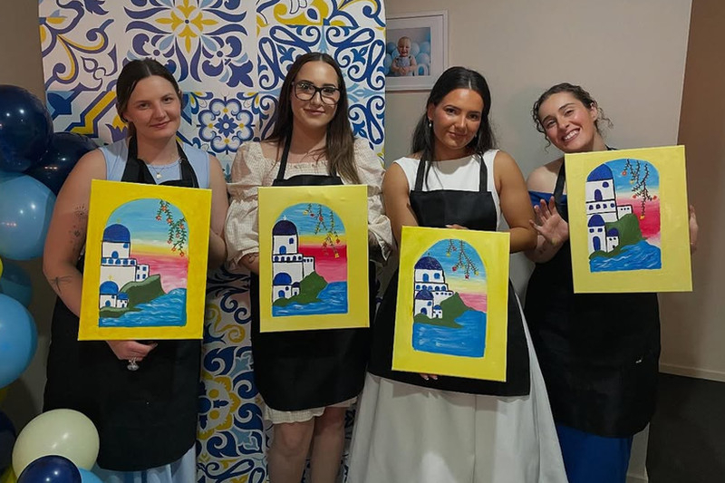 Four friends in aprons holding up their painted canvases after a paint class