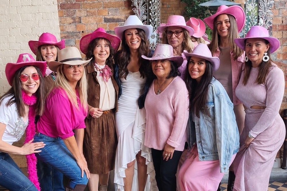 Friends of every age in pink cowgirl hats and pink, denim and white outfits around the bride in a white dress in a brick courtyard