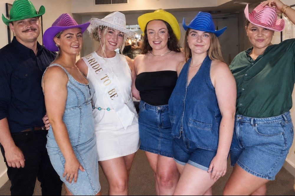 A bride to be in white with a white hat and sash beside friends in denim and cowgirl hats in green, purple, yellow, blue and pink sequins