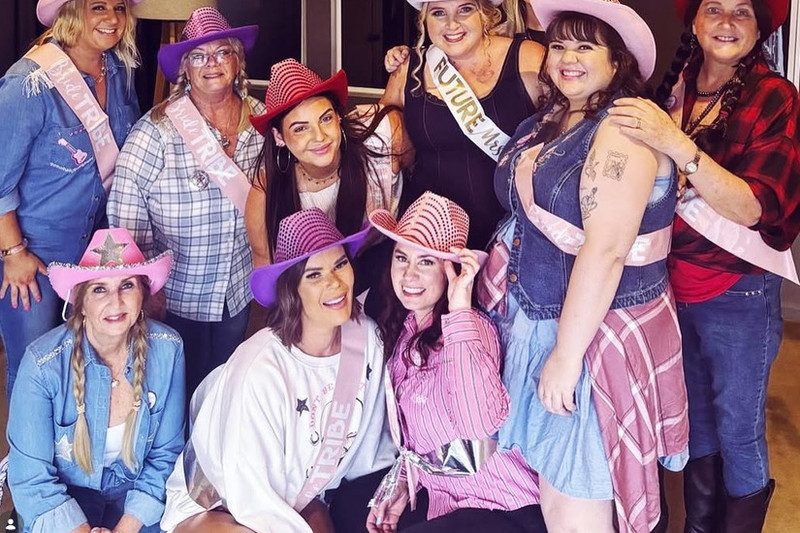 Friends in pink and purple cowgirl hats, plaid shirts, denim and sashes crowded around the bride