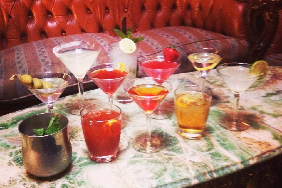 Colourful cocktails lined up on a marble table in front of a red leather sofa