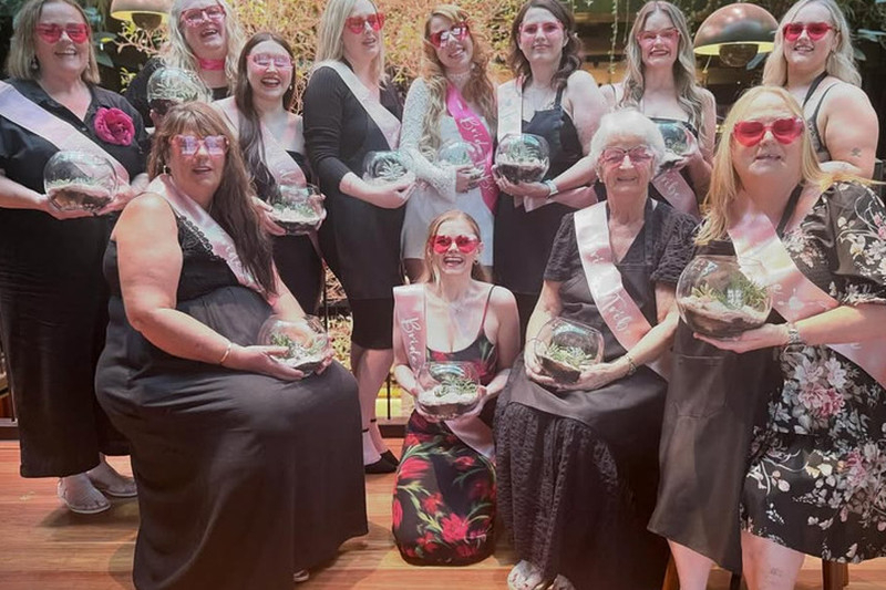 A hens group of all ages in pink heart sunglasses and sashes holding up the terrariums they made