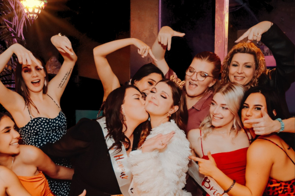 Friends dancing and pointing at the bride in white while one kisses her cheek