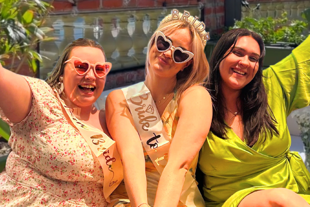 A bride in a sash and tiara laughing with two friends on a rooftop
