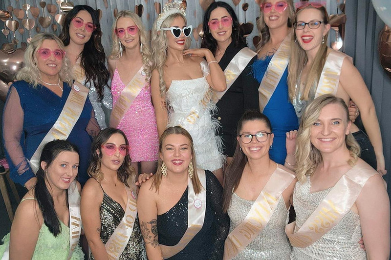 Hens in sashes and pink sunglasses gathered round the bride in front of rose gold hearts