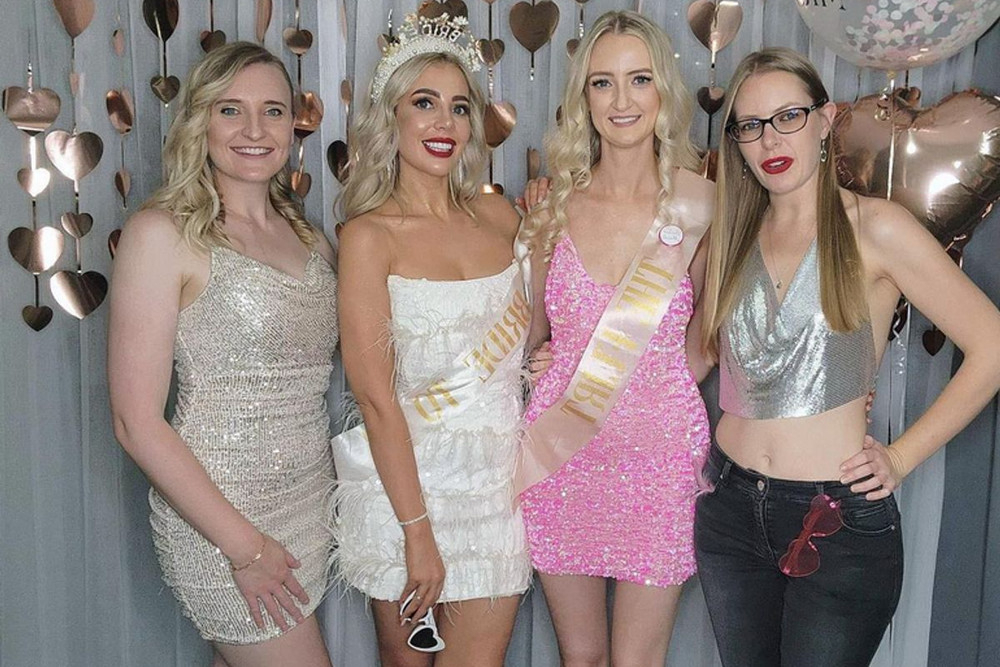 Four friends dressed up for the night, the bride in a tiara and sash, in front of a wall of rose gold hearts