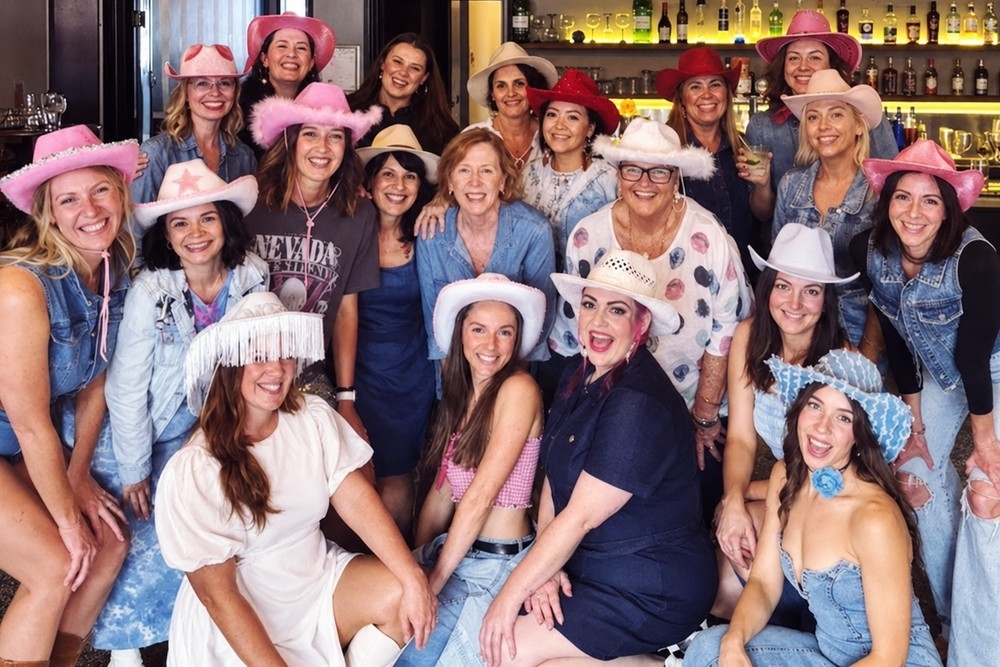 A big group of friends of every age in denim, fringe and pink and white cowgirl hats crowded together at a bar