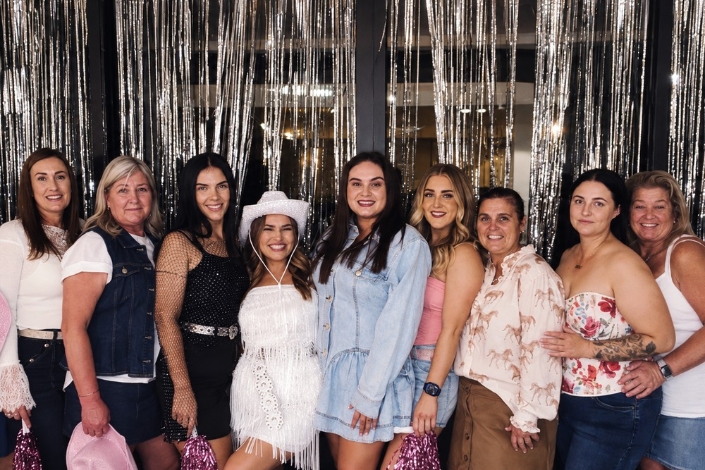 A bride in a white fringe mini and white cowgirl hat with friends of every age in denim jackets, florals and boots in front of a silver tinsel curtain