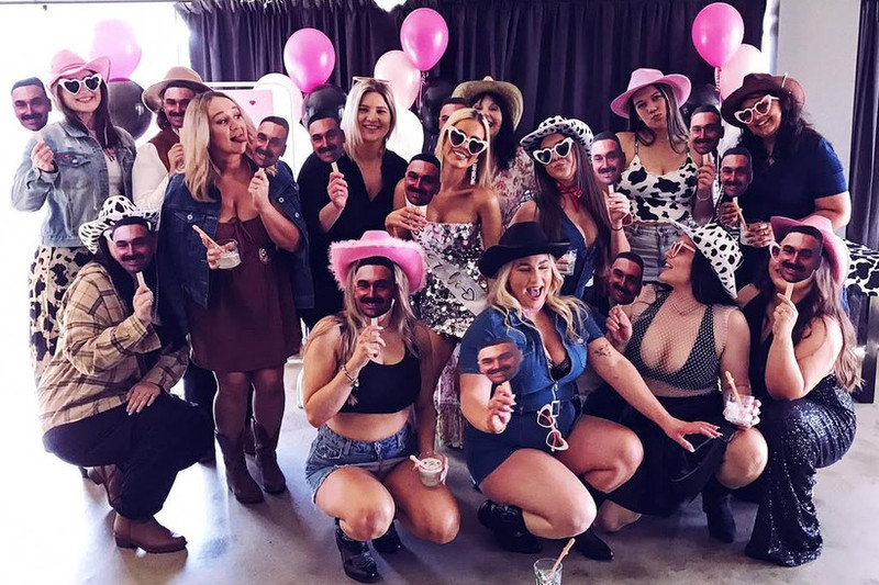 A cowgirl hens group in pink hats, cow print and denim holding up masks of the groom’s face around the bride in a sequin dress