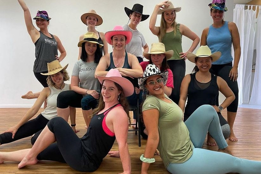 A hens crew in cowboy hats and activewear posing together on the floor of a bright pole studio